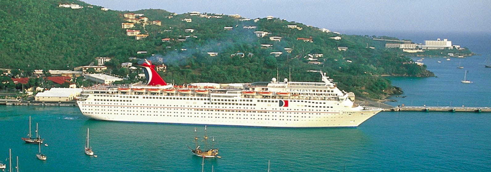 Fartyget Carnival Ecstasy ligger vid en hamn med en grön kulle i bakgrunden och mindre segelbåtar i framkant.