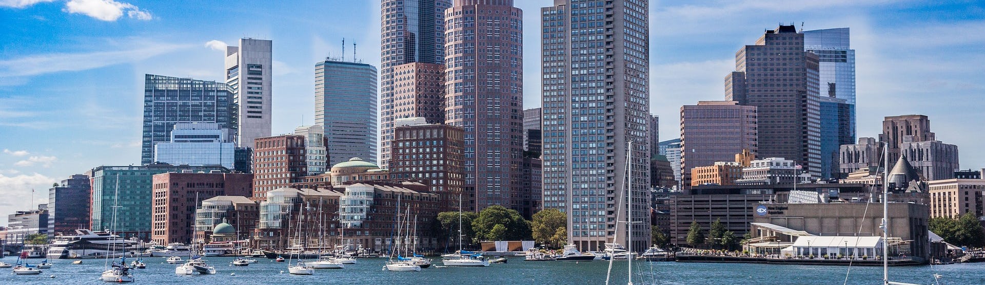 Bild på Bostons skyline med havet framför och flera segelbåtar.