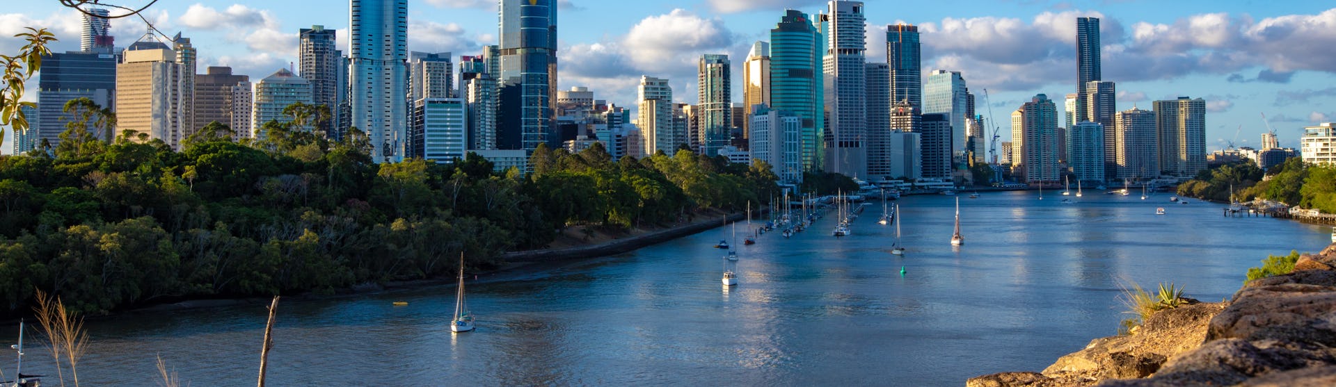 Bild på Brisbane med segelbåtar i vattnet, höghus och vacker natur.