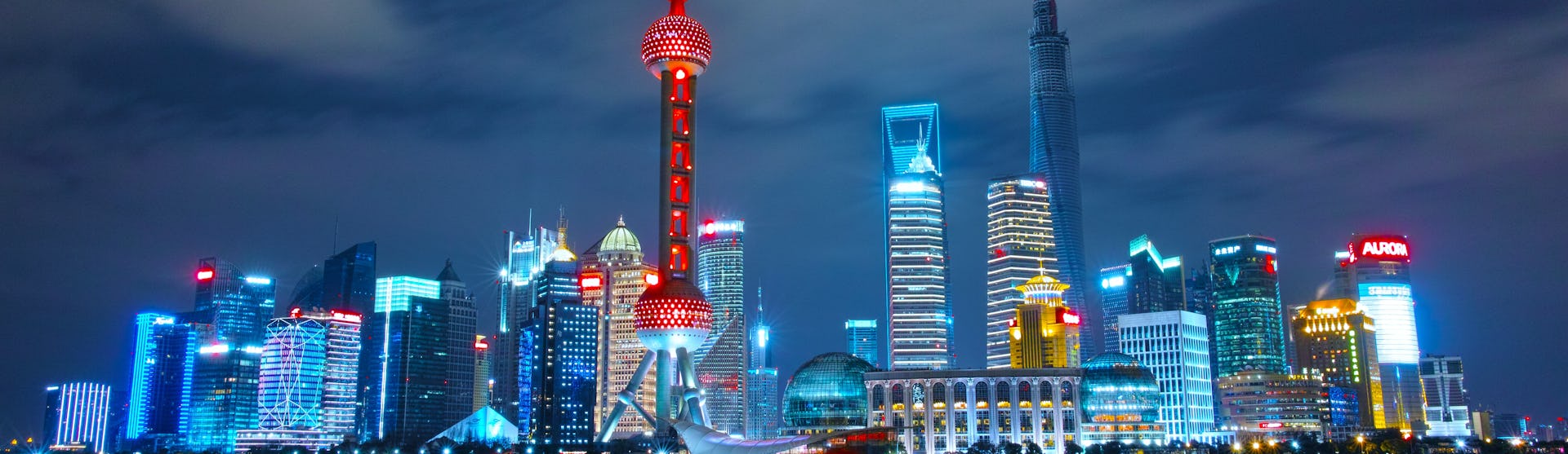 Bild tagen från vattnet på Shanghai's skyskrapor i kvällsljus. Byggnaderna lyser i olika färger.