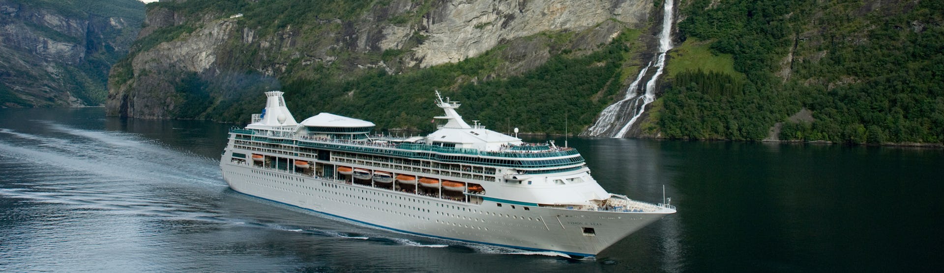 Vision of the Seas kryssar genom de storslagna norska fjordarna. I bakgrunden syns gröna berg och ett vattenfall.