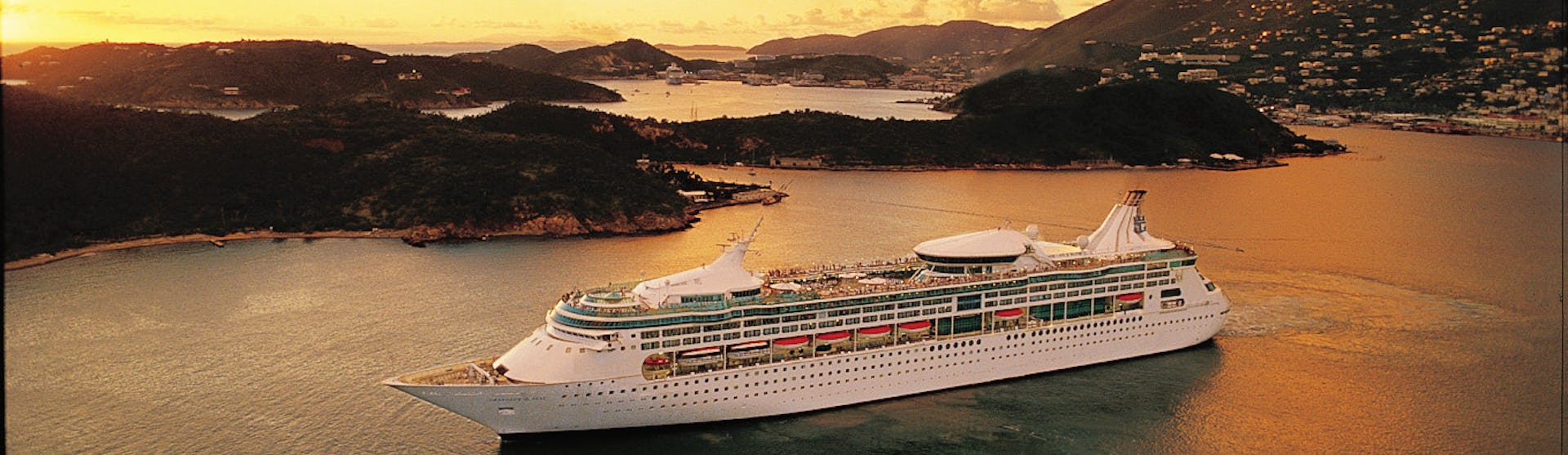 Bild på Rhapsody of the Seas, taget från sidan, utanför en Karibisk ö i solnedgången.