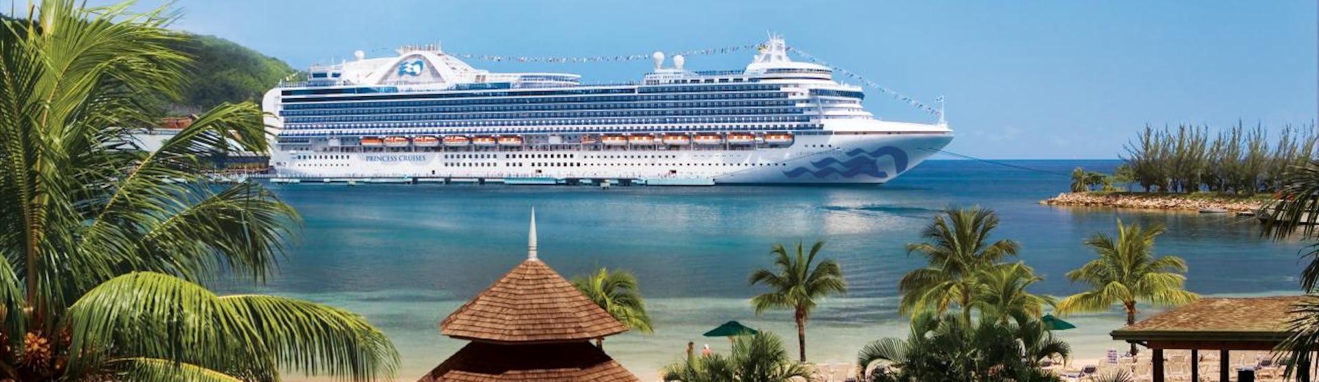 Fartyget Crown Princess ligger ankrad utanför en hamn i exotiska Jamaica. I framkant syns palmer och vit sand.